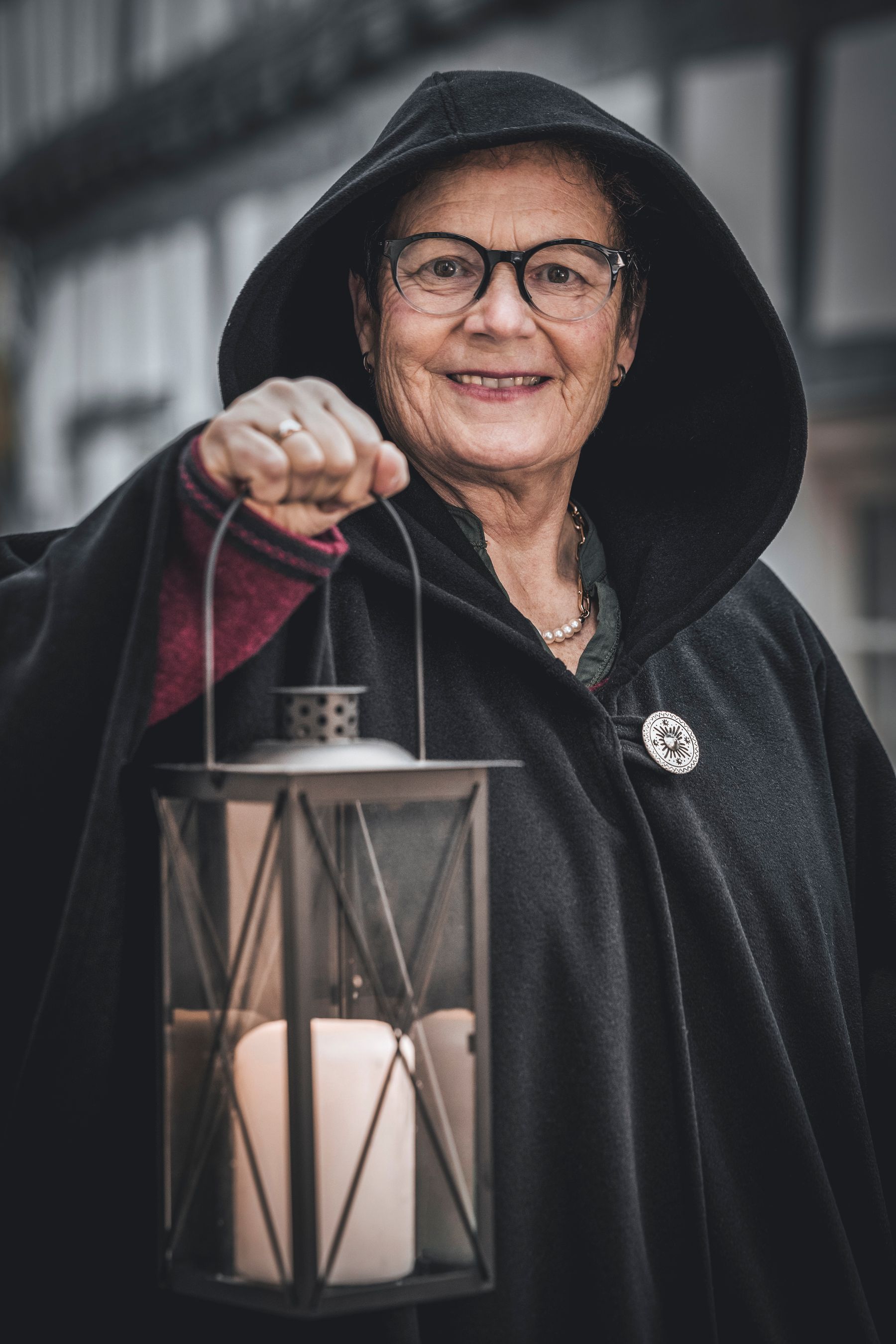 An older person wearing glasses and a dark hooded cloak smiles while holding a lantern with a lit candle inside, standing outdoors in front of a blurred building.